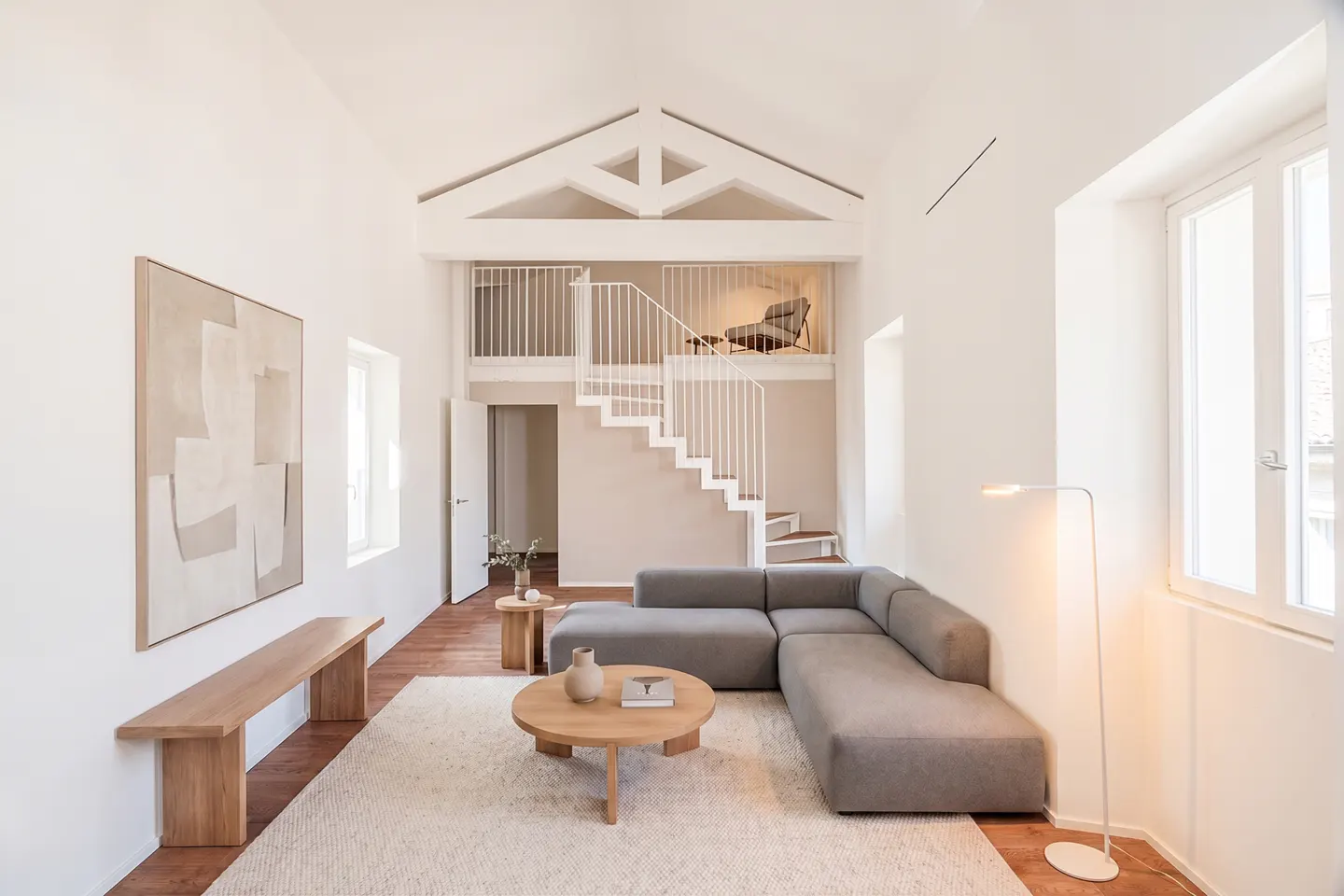 Bright, modern living room with a gray sectional sofa, round wood table, and white staircase leading to a loft area. Abstract art hangs on the white wall.