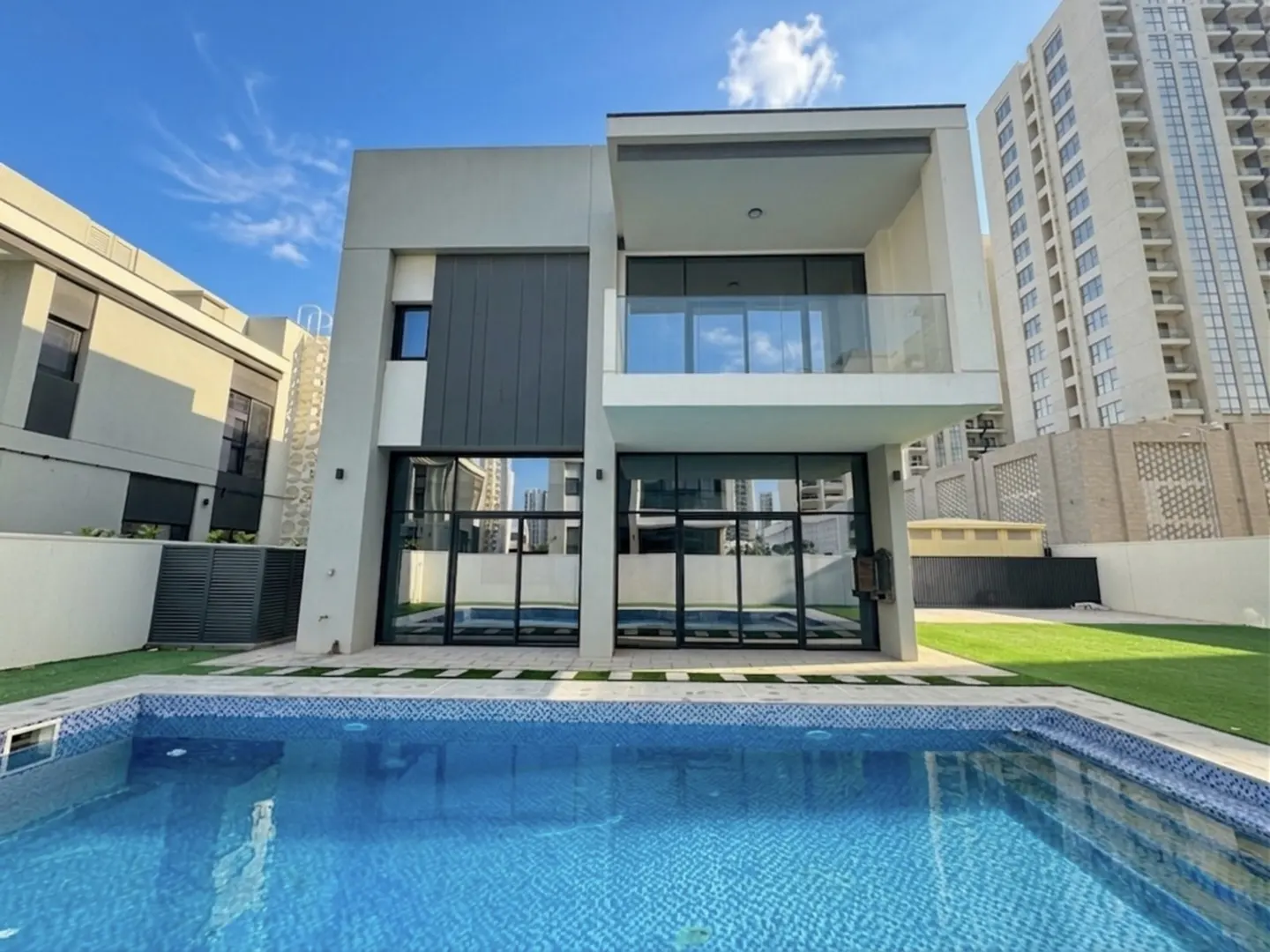 Modern two-story house with a blue tiled swimming pool in the foreground. Large glass doors and a balcony are visible.