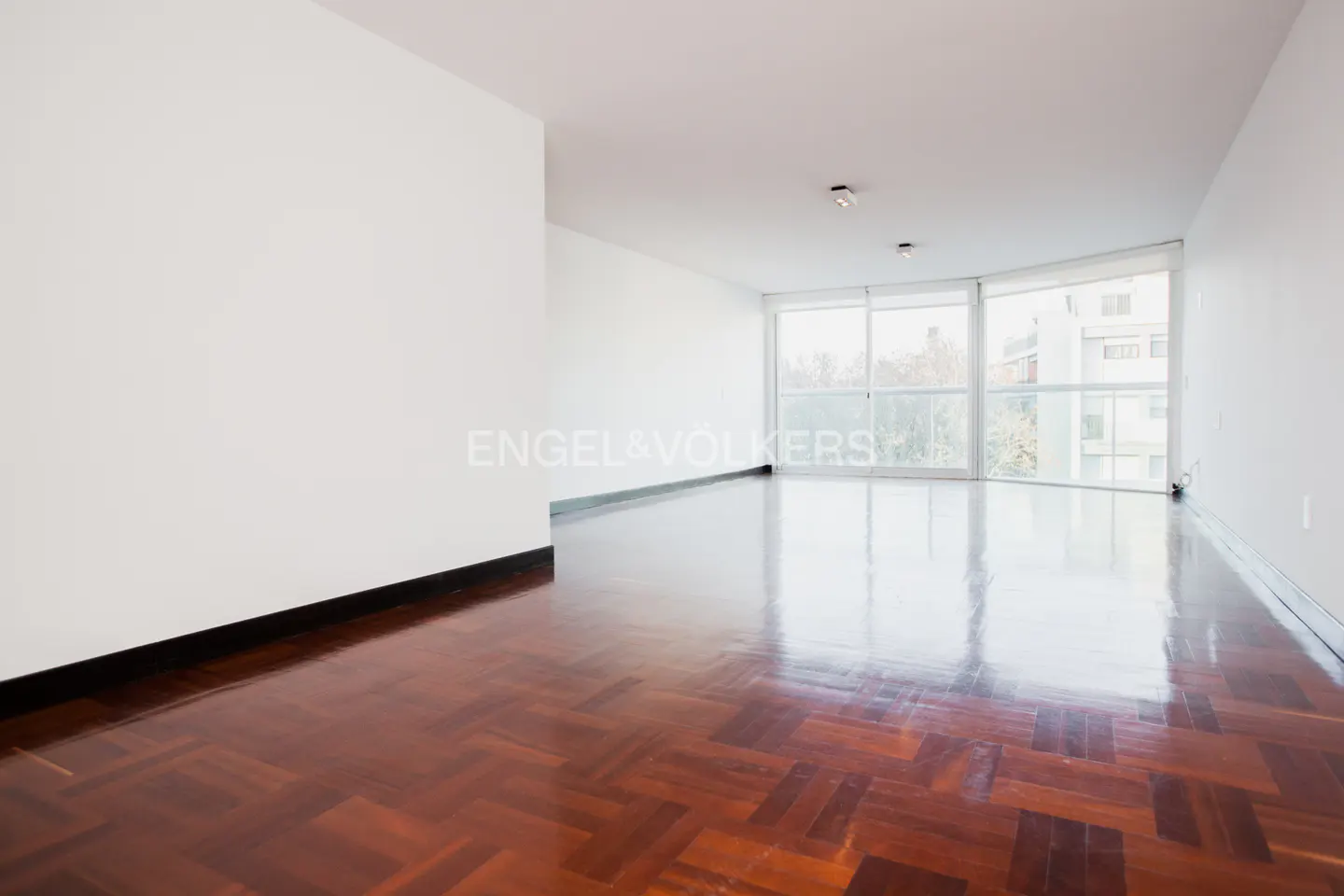 Empty room with herringbone wood floors, white walls, and large windows overlooking trees and buildings.