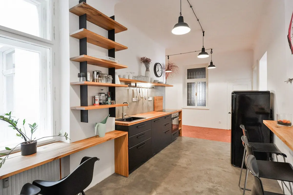 A modern kitchen with black cabinets, wood countertops, and open shelving. A black refrigerator and bar stools are visible.