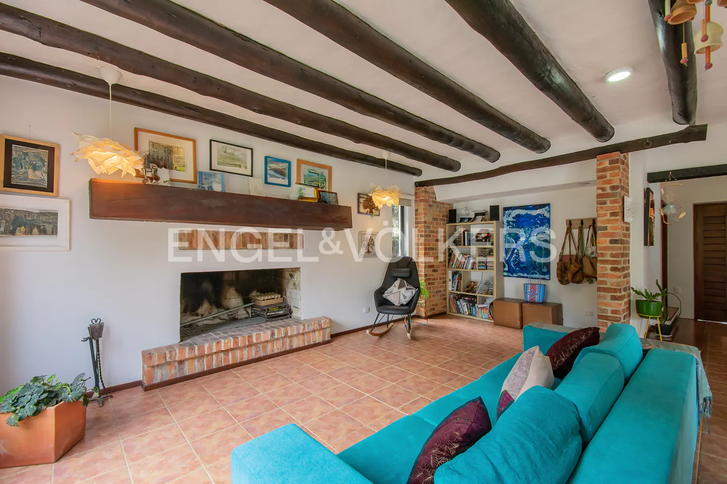 Bright living room with a turquoise sofa, brick fireplace, and dark wood beams on the ceiling. Bookshelves and artwork add to the cozy atmosphere.