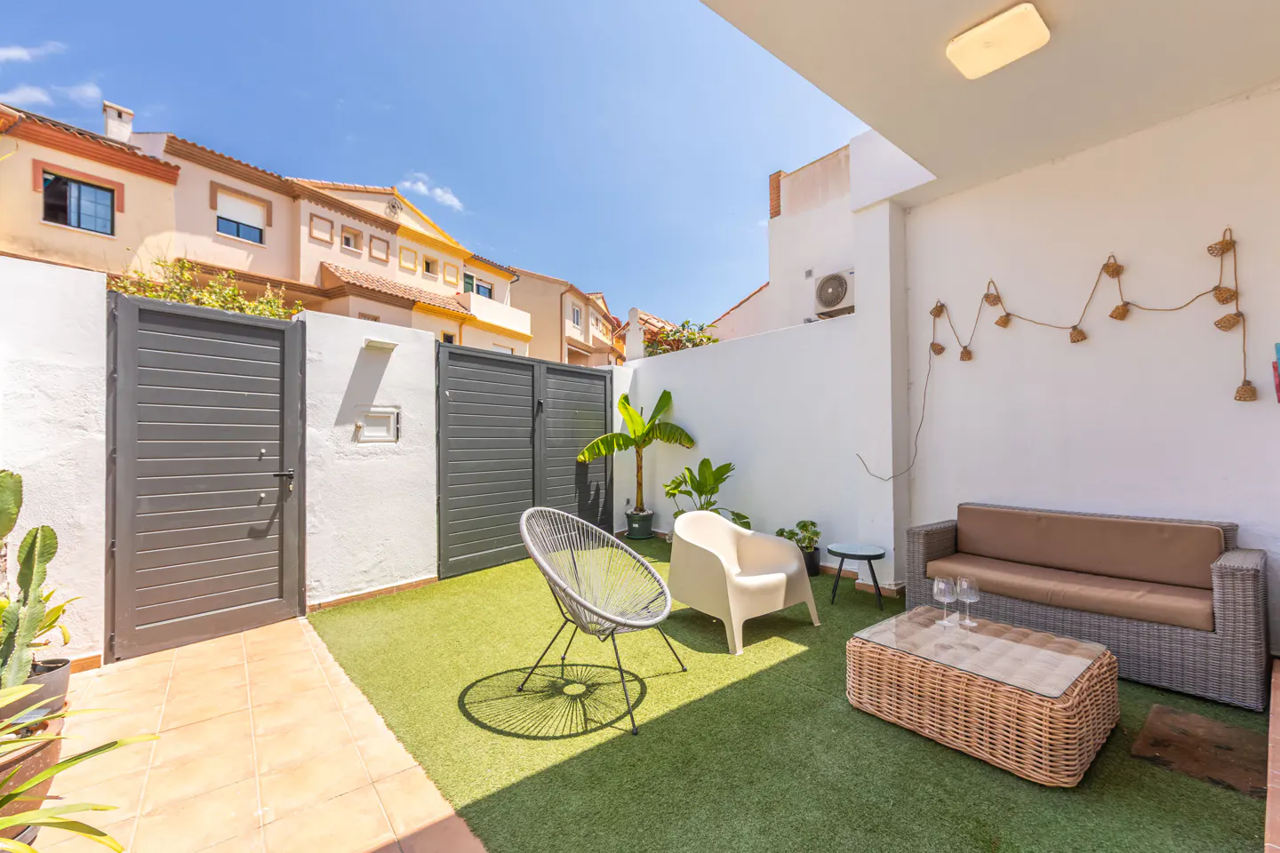 A cozy outdoor patio with a gray sofa, wicker table, and chairs on artificial grass. A white wall is decorated with string lights.