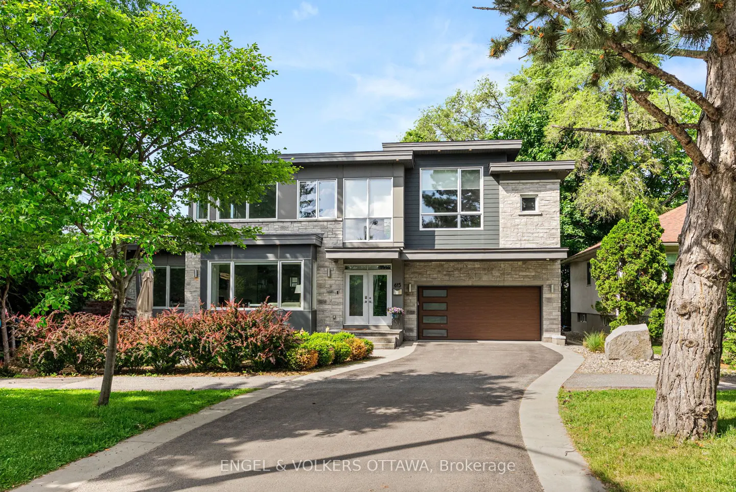 Two-story modern home with gray siding, stone accents, and a brown garage door. A curved driveway leads to the house, surrounded by green trees.