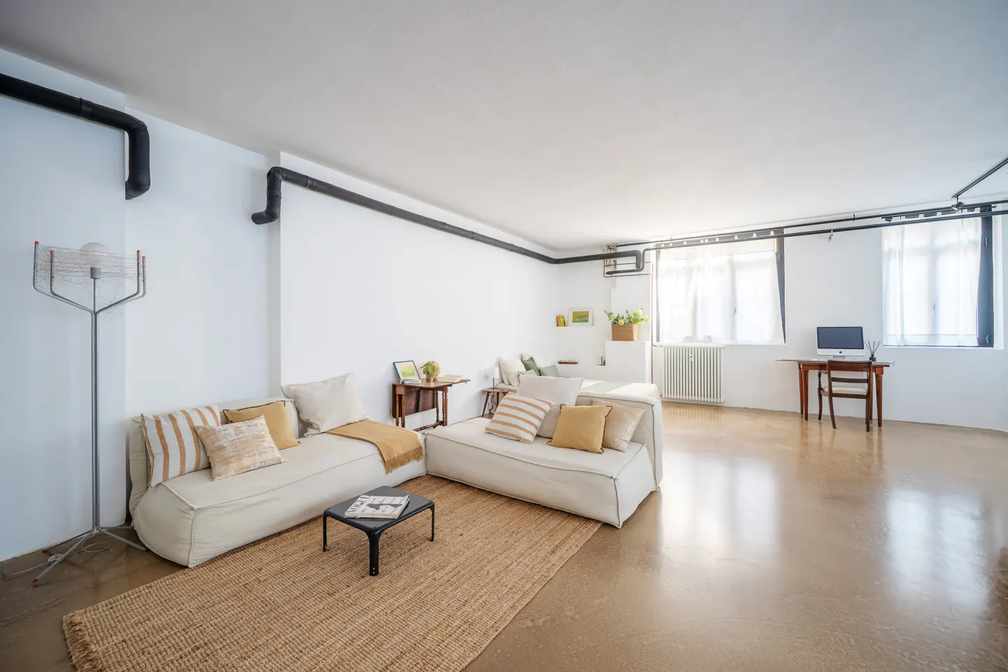 Bright, modern living room with white walls, beige couches, and a jute rug. Black pipes run along the ceiling. A desk sits by the window.