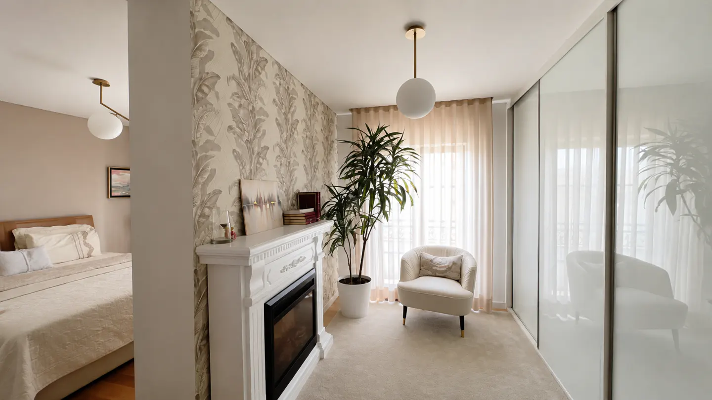 Bedroom with a white fireplace, chair, and plant. A bed is visible through a doorway. Wallpaper with leaf pattern.