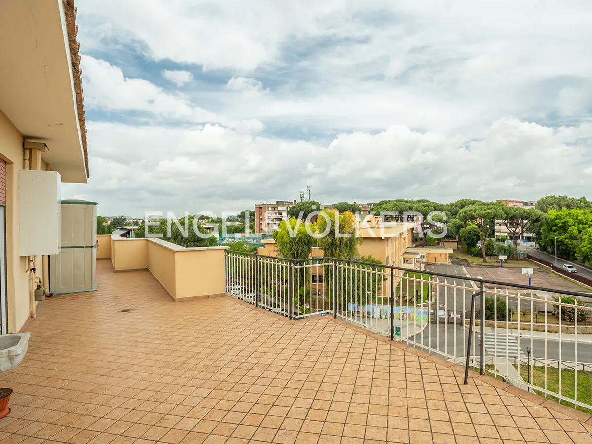 Tiled rooftop terrace with white railings overlooks a cityscape with trees and buildings under a cloudy sky.