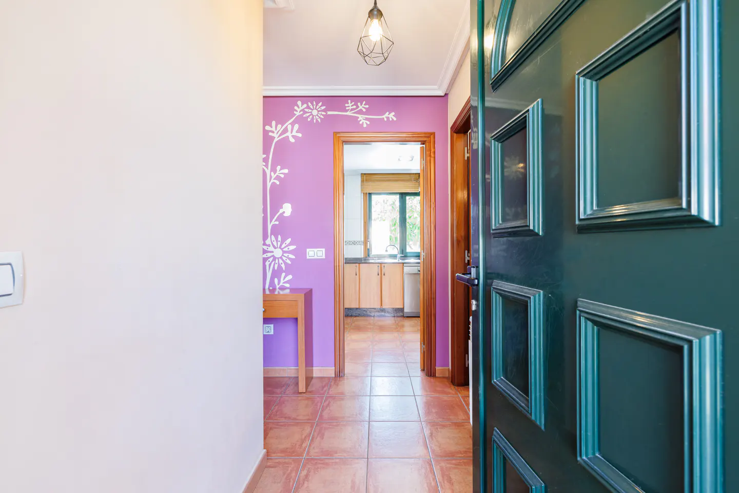 Open teal door reveals a hallway with terracotta tile floor, leading to a kitchen. A purple wall with white floral design is visible.