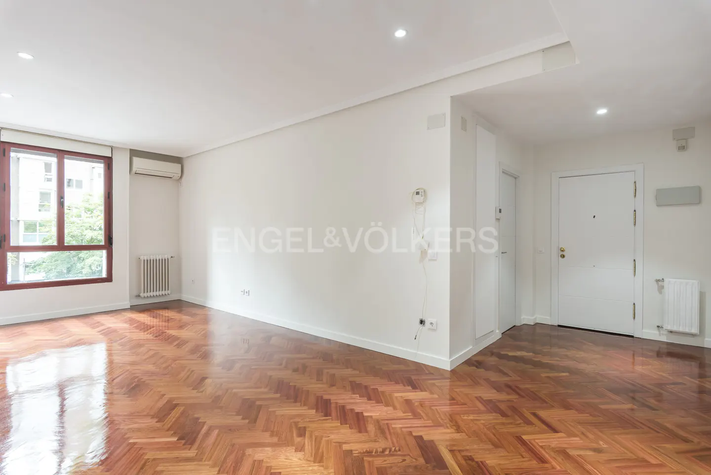 Bright, empty room with herringbone wood floors, white walls, and a window with a brown frame. Engel & Völkers logo on the wall.