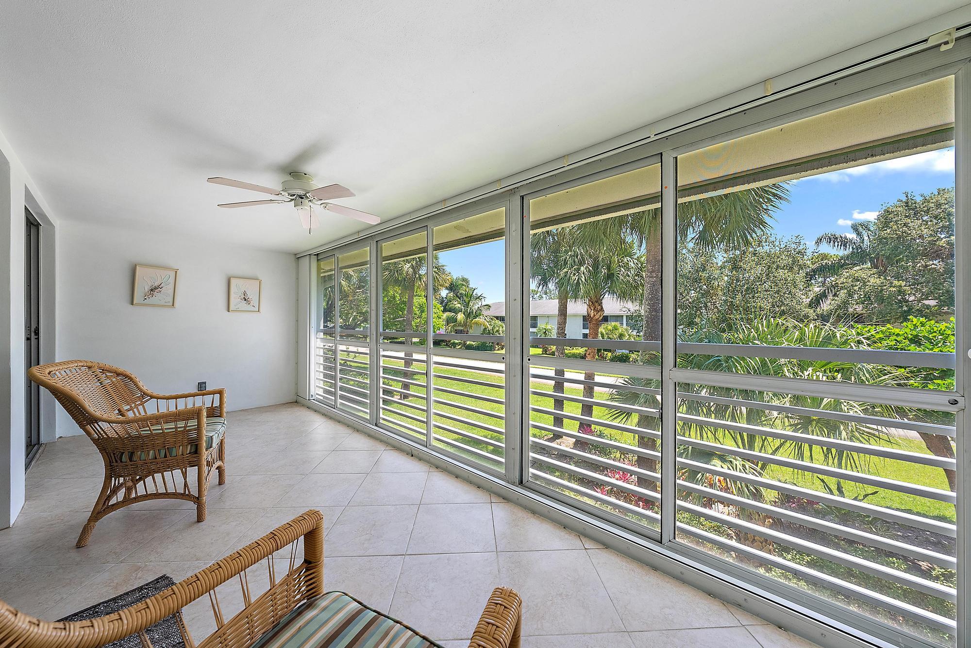Sunroom with two wicker chairs, tile floors, ceiling fan, and large windows overlooking a lush, green tropical yard.