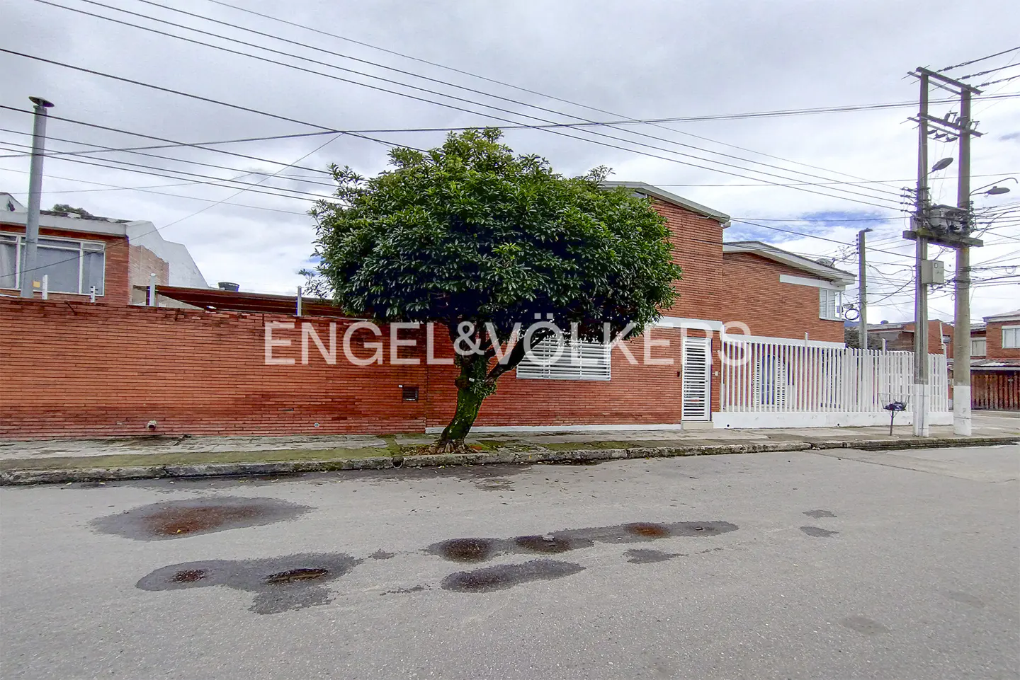 Exterior view of a red brick house with a white fence and a tree in front. The Engel & Völkers logo is visible on the wall.