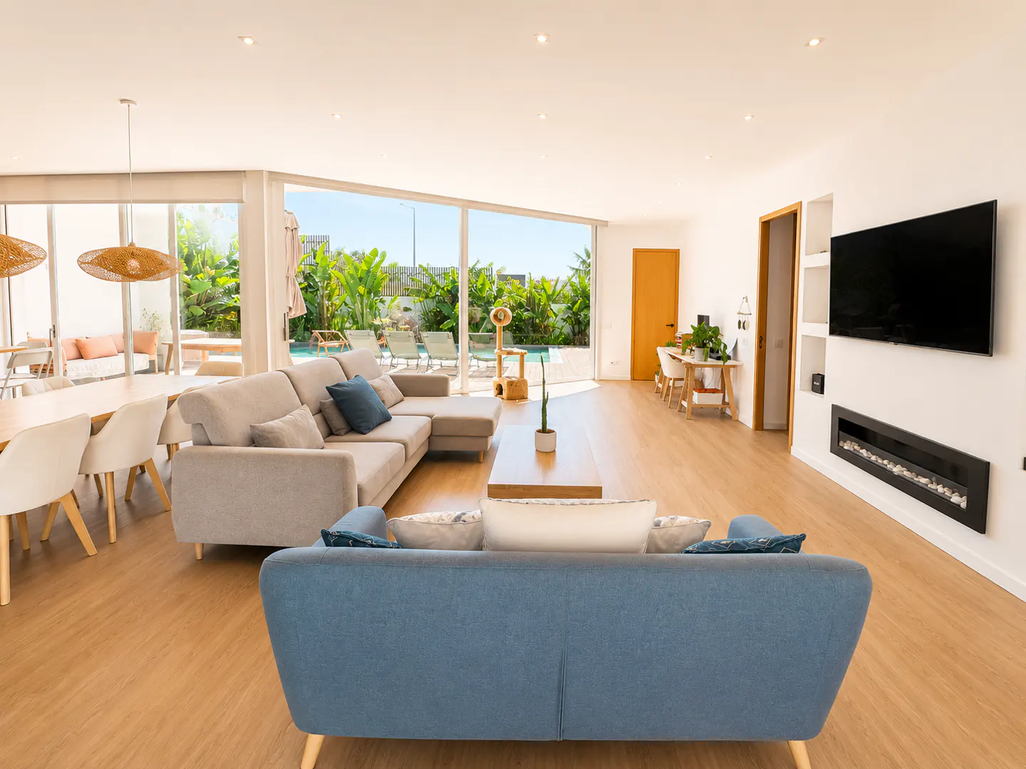 Modern open-plan living room with light wood floors, two sofas, a dining area, and a view of a pool through glass walls.