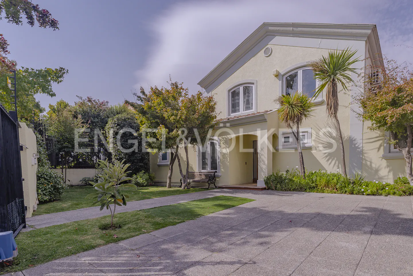 Two-story yellow house with white trim, a concrete driveway, and green lawn with trees. Engel & Volkers logo is visible.