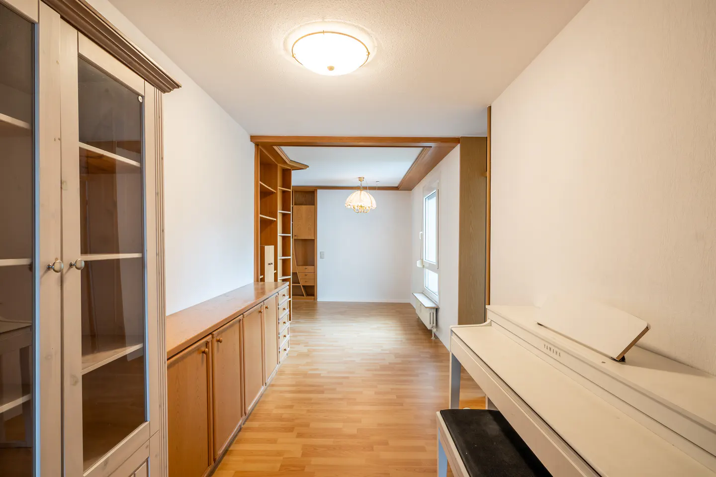 A bright room with wood floors, built-in cabinets, a white piano, and a view into another room.