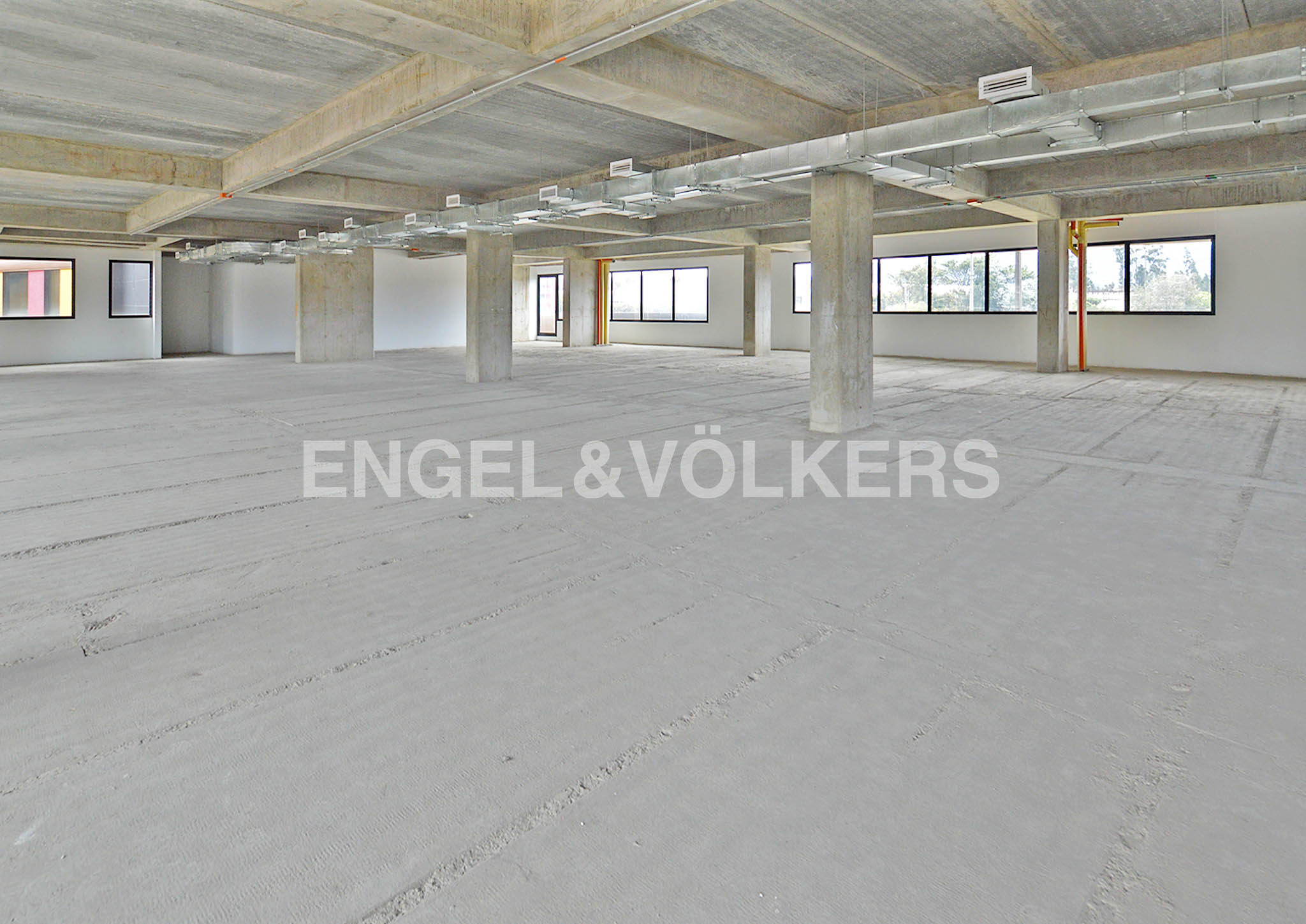 An empty commercial space with concrete floors, columns, and ceiling. Windows line the far wall, letting in natural light. The Engel & Völkers logo is in the foreground.