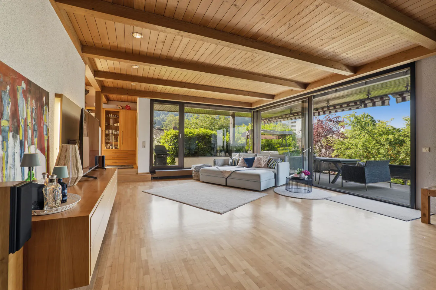Modern living room with wood-beamed ceiling, light wood floors, large windows, and a view of a patio with outdoor seating.