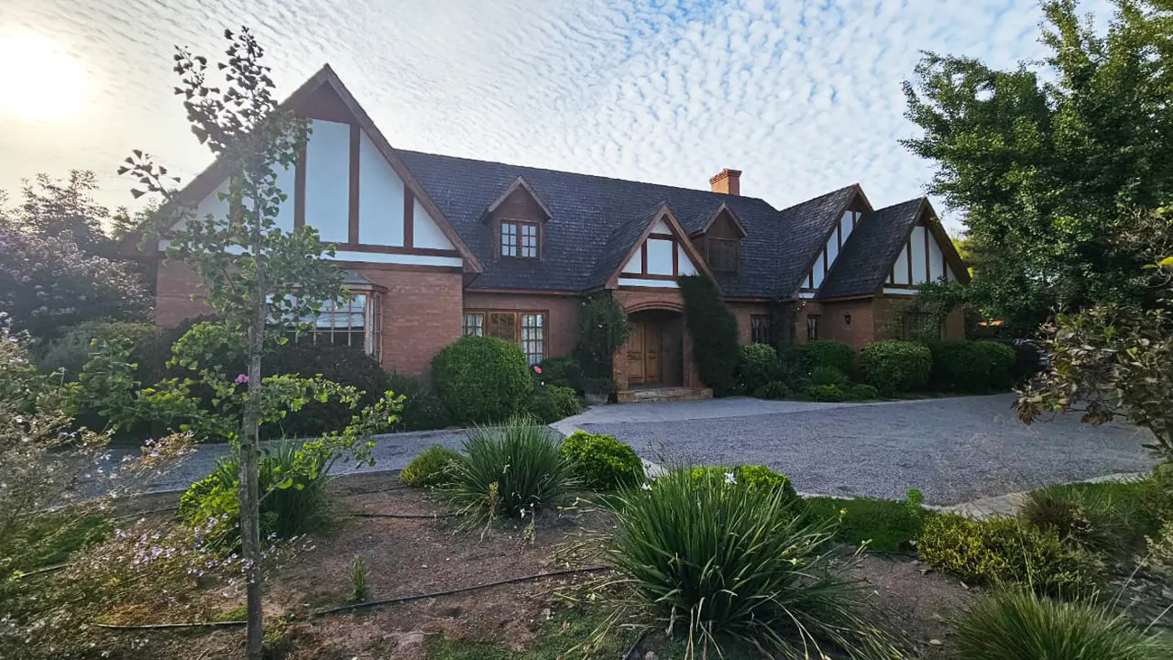 A red brick Tudor-style house with a dark roof and white gables, surrounded by green landscaping and a gravel driveway.