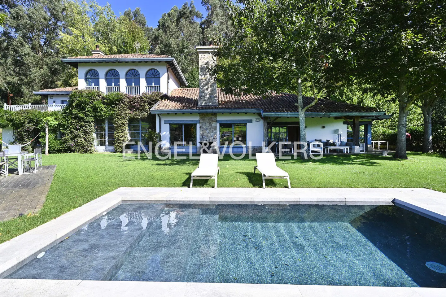 A white house with a red tile roof overlooks a pool with two lounge chairs on the lawn.