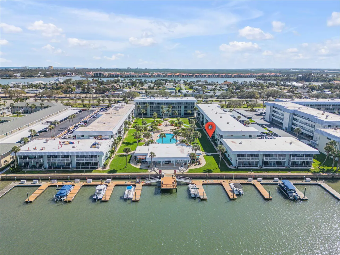 Aerial view of a waterfront condo complex with a pool, boat docks, and a red location pin on one of the buildings.