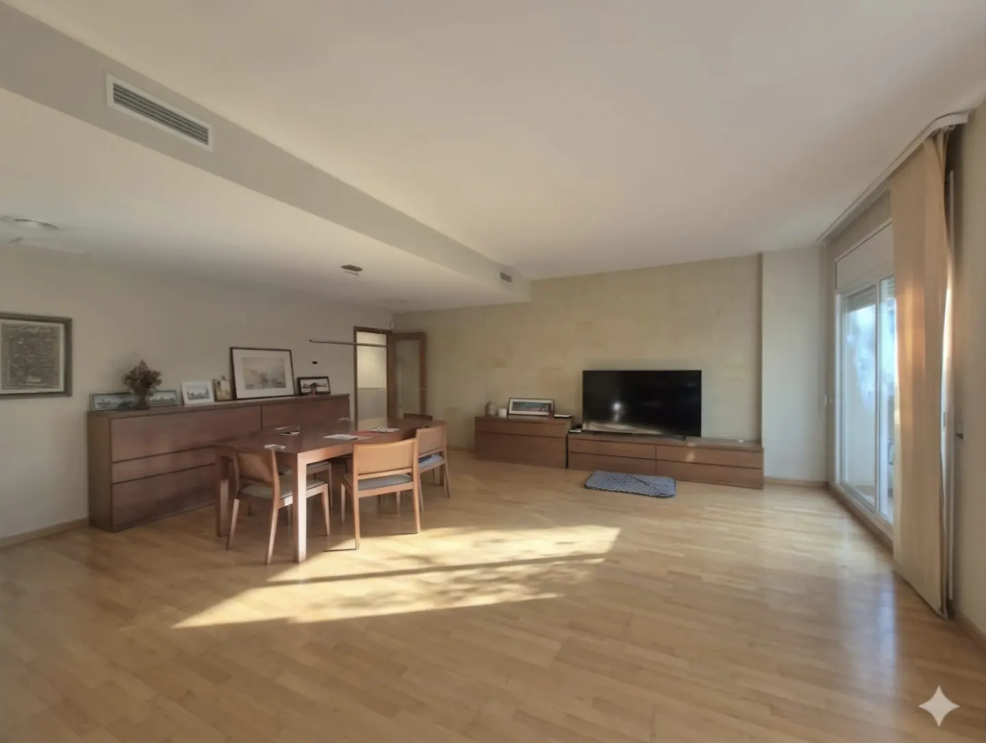 A bright, spacious living room with wood floors, a dining table, and a large TV. Sunlight streams in from a balcony door.