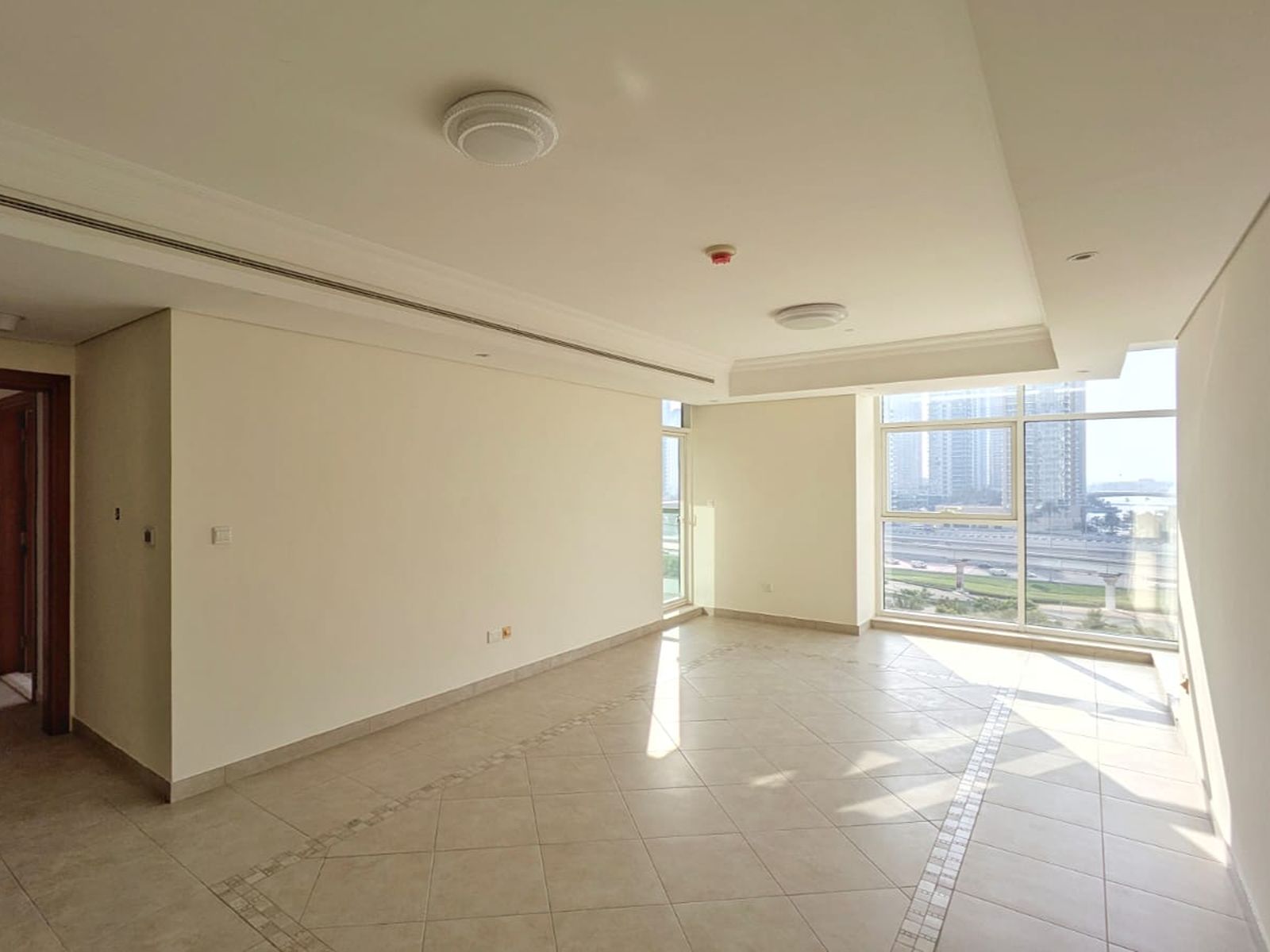 Empty, bright living room with beige tiled floors, cream walls, and large windows overlooking a city highway.