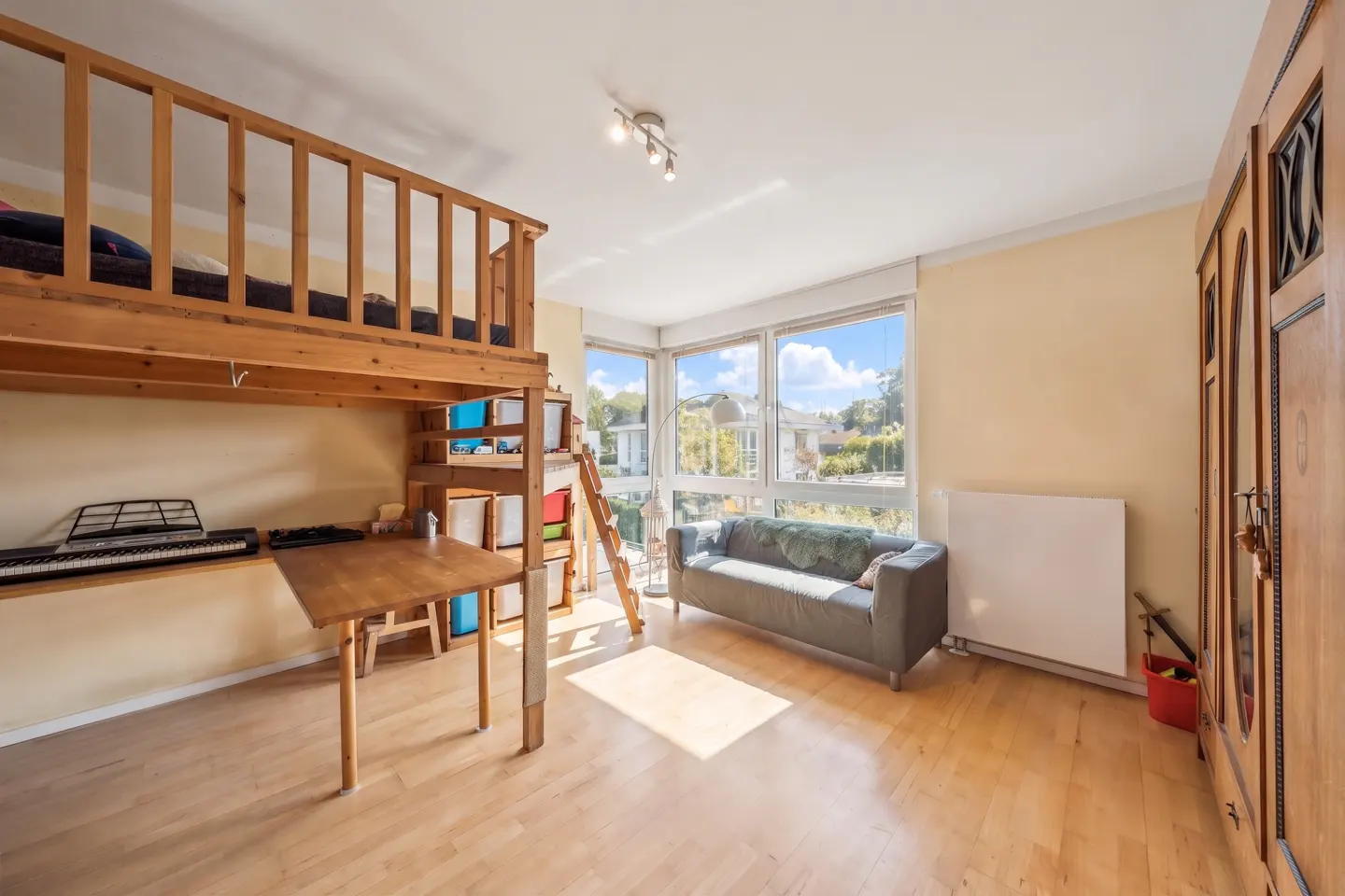 Bright room with a wooden loft bed above a desk, a grey sofa near windows, and a wooden wardrobe.