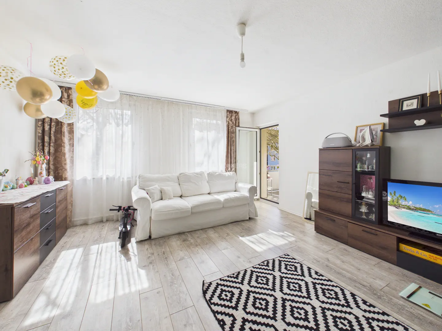 Bright living room with white walls, wood floors, and a white sofa. Balloons hang from the ceiling. A TV and cabinet are on the right.