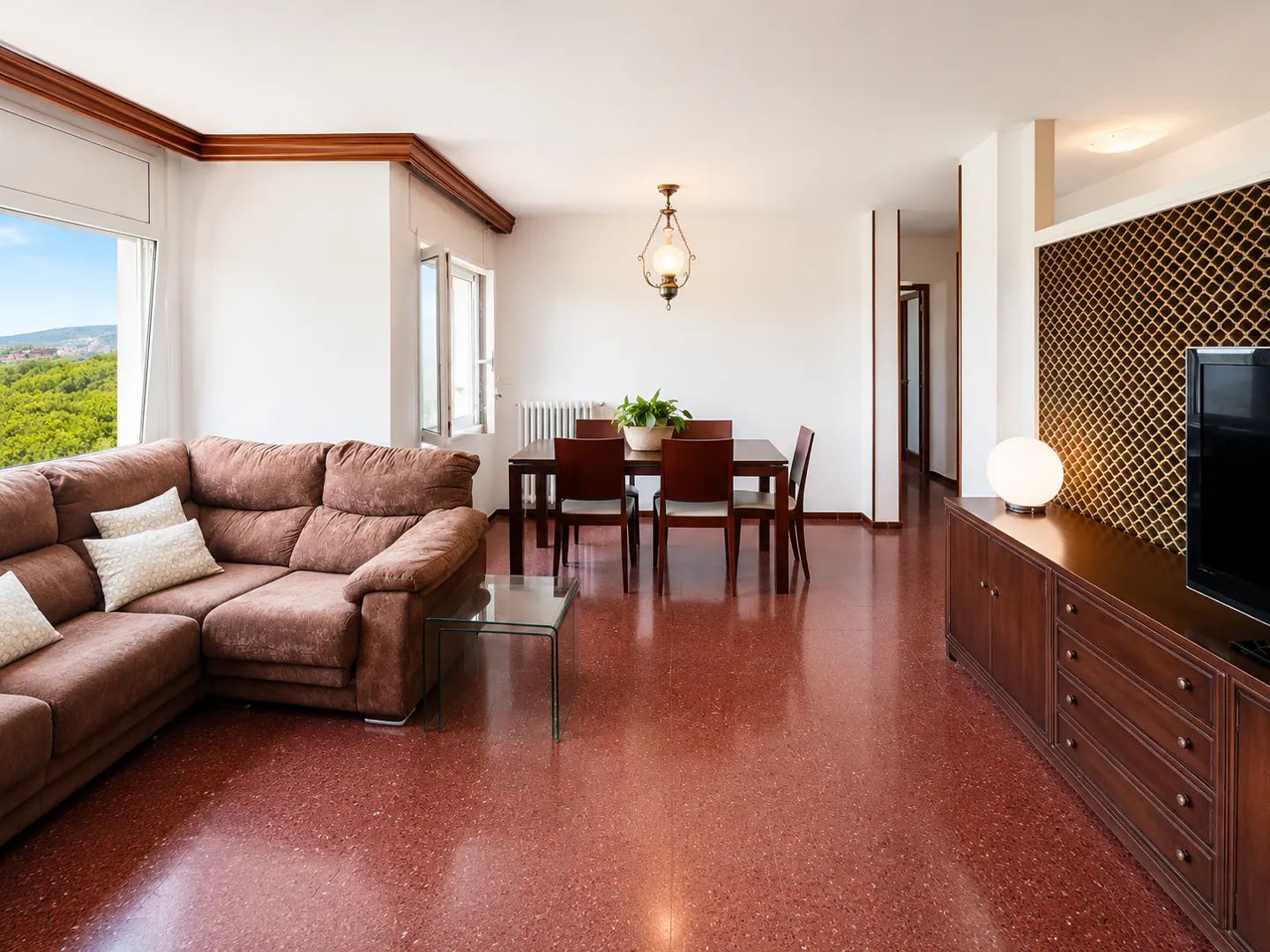 Spacious living room with a brown sectional sofa, dining area, red terrazzo floors, and a view of trees through the window.
