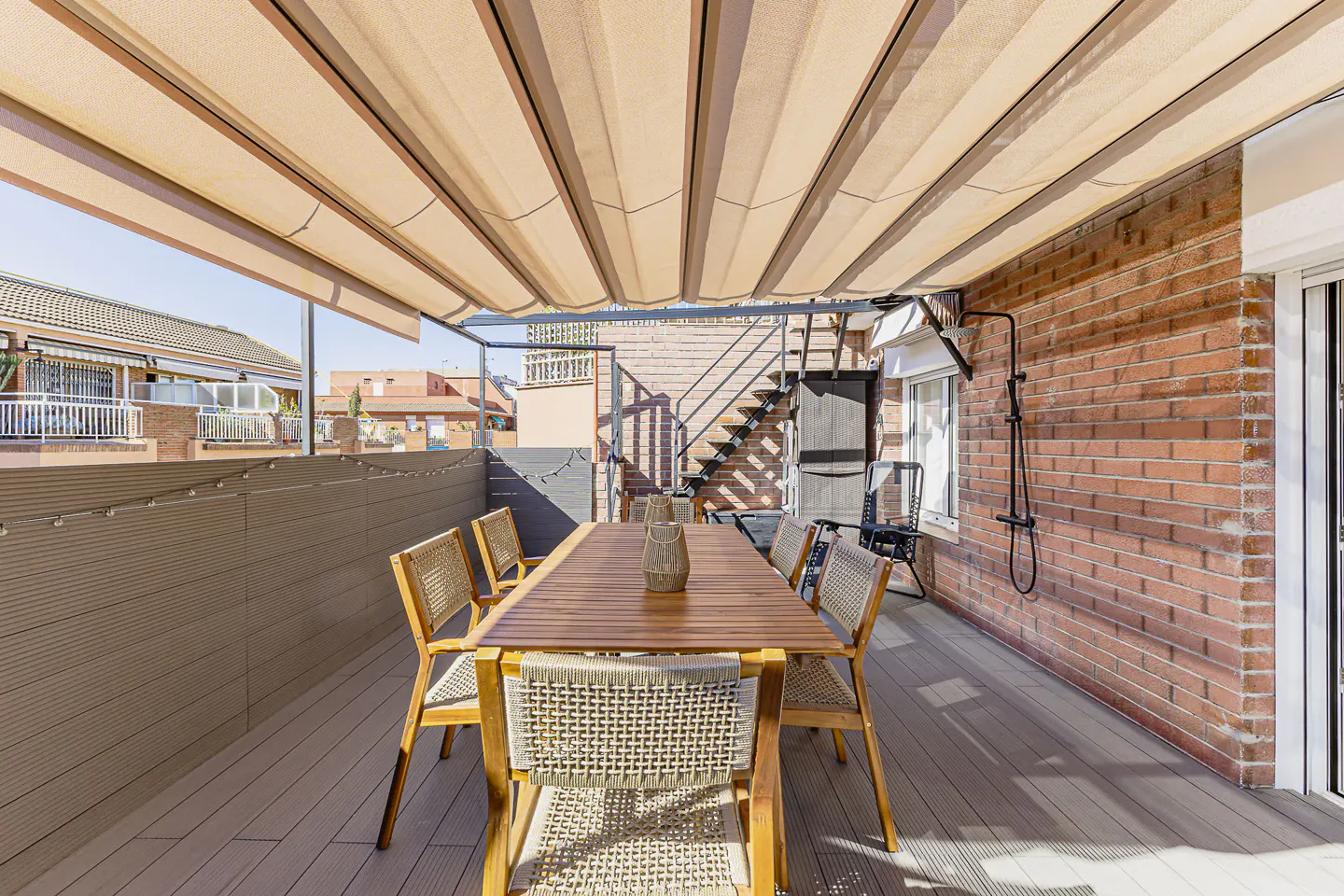 Outdoor patio with a wooden table, chairs, and a retractable awning. Brick wall and stairs in the background.
