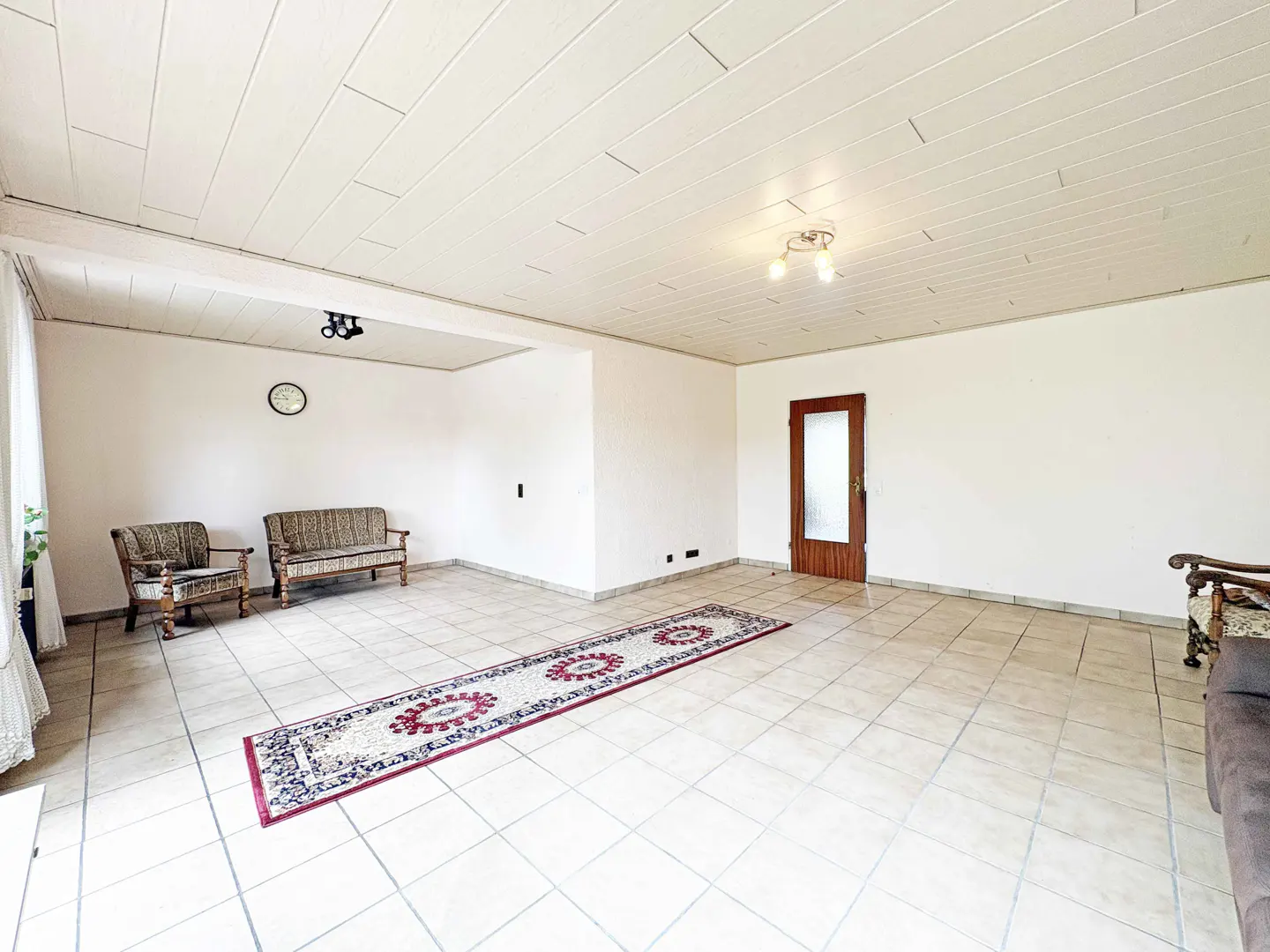 Bright, empty living room with tile floor, white walls, and a long, patterned rug. Two sofas and a chair are visible.