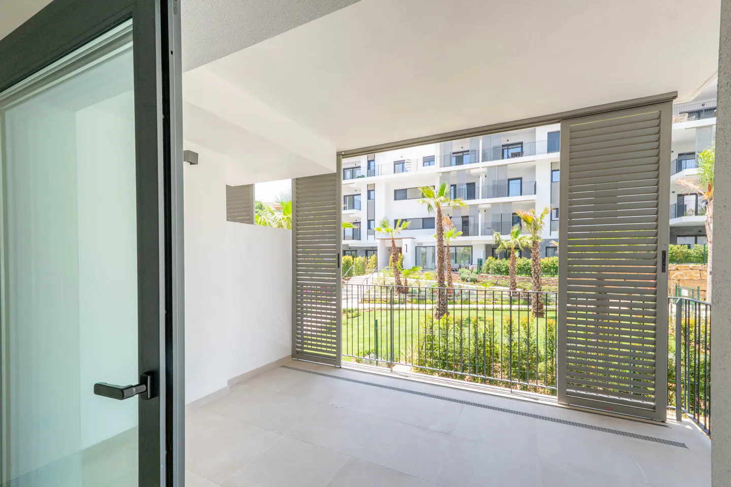 View from a modern apartment balcony with grey shutters looking out onto a landscaped courtyard with palm trees.
