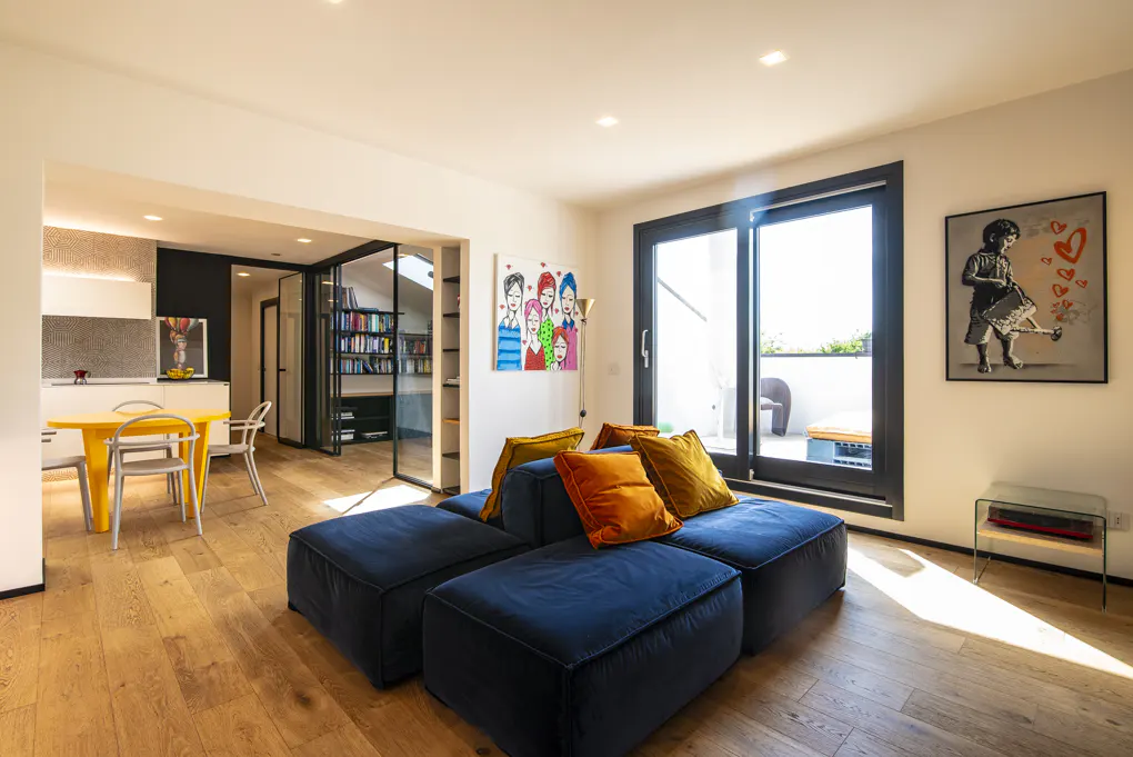 Bright living room with hardwood floors, a blue modular sofa with orange pillows, and a yellow table with chairs. Artwork adorns the walls.