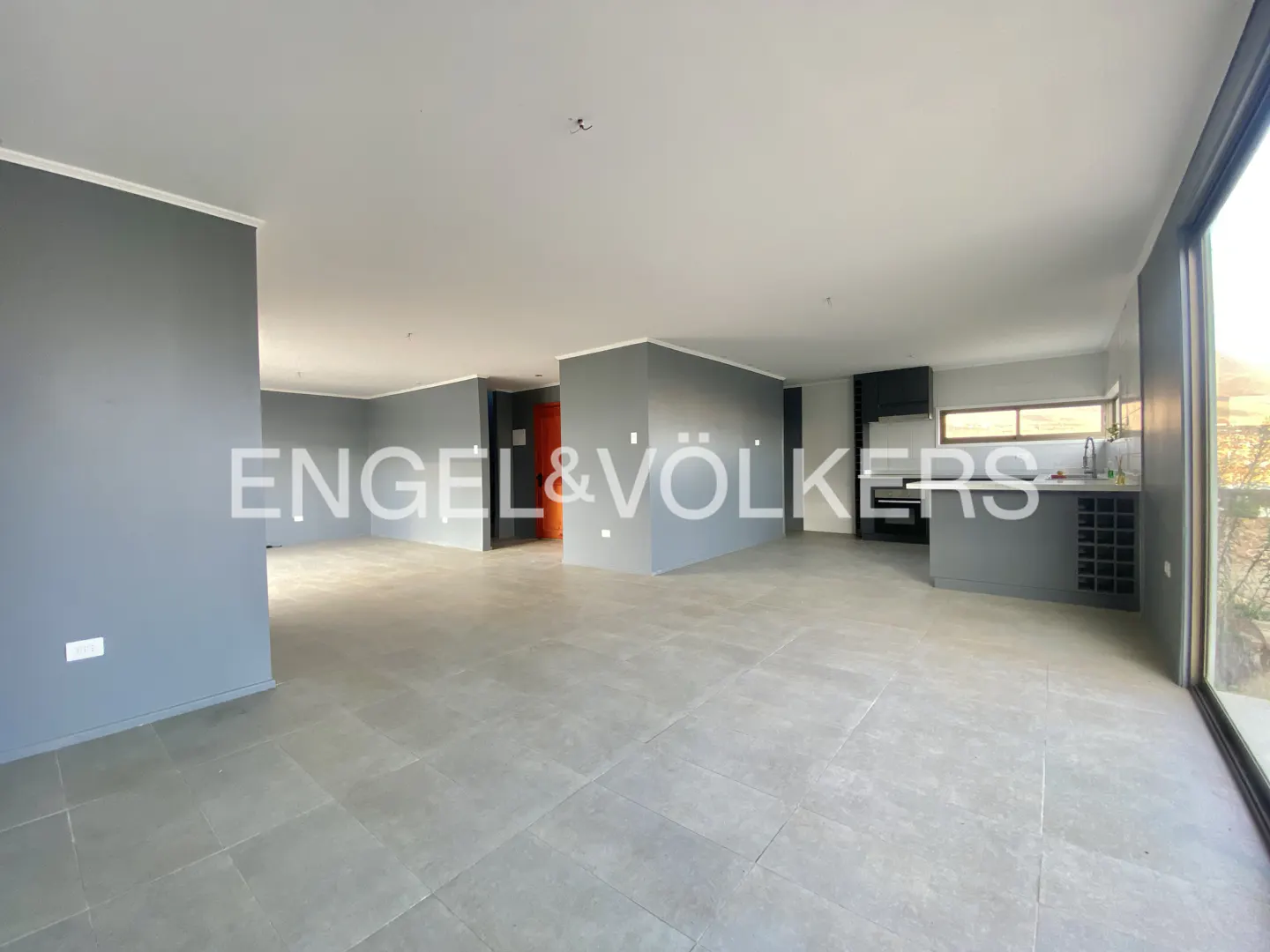 An open-concept living space with gray walls, tile floors, and a modern kitchen area. Large windows offer natural light. Engel & Völkers logo is visible.