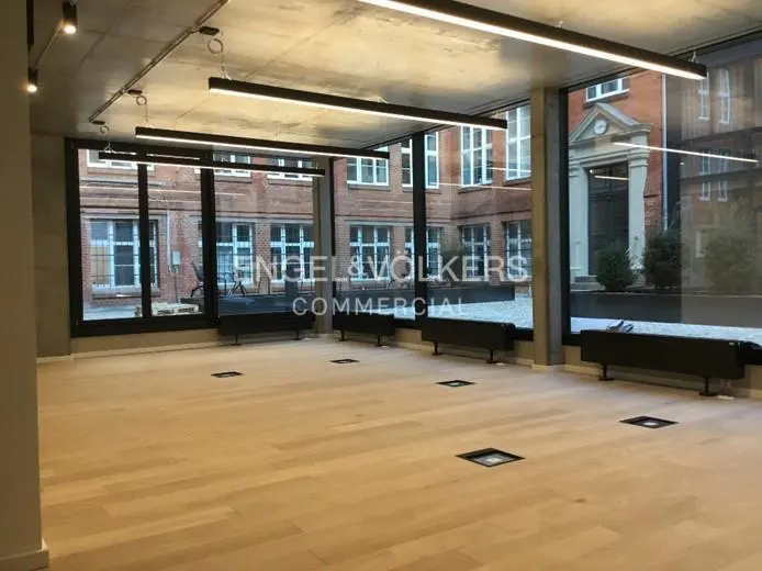 Bright, empty commercial space with light wood floors, black window frames, and concrete ceiling. Engel & Volkers logo on the window.