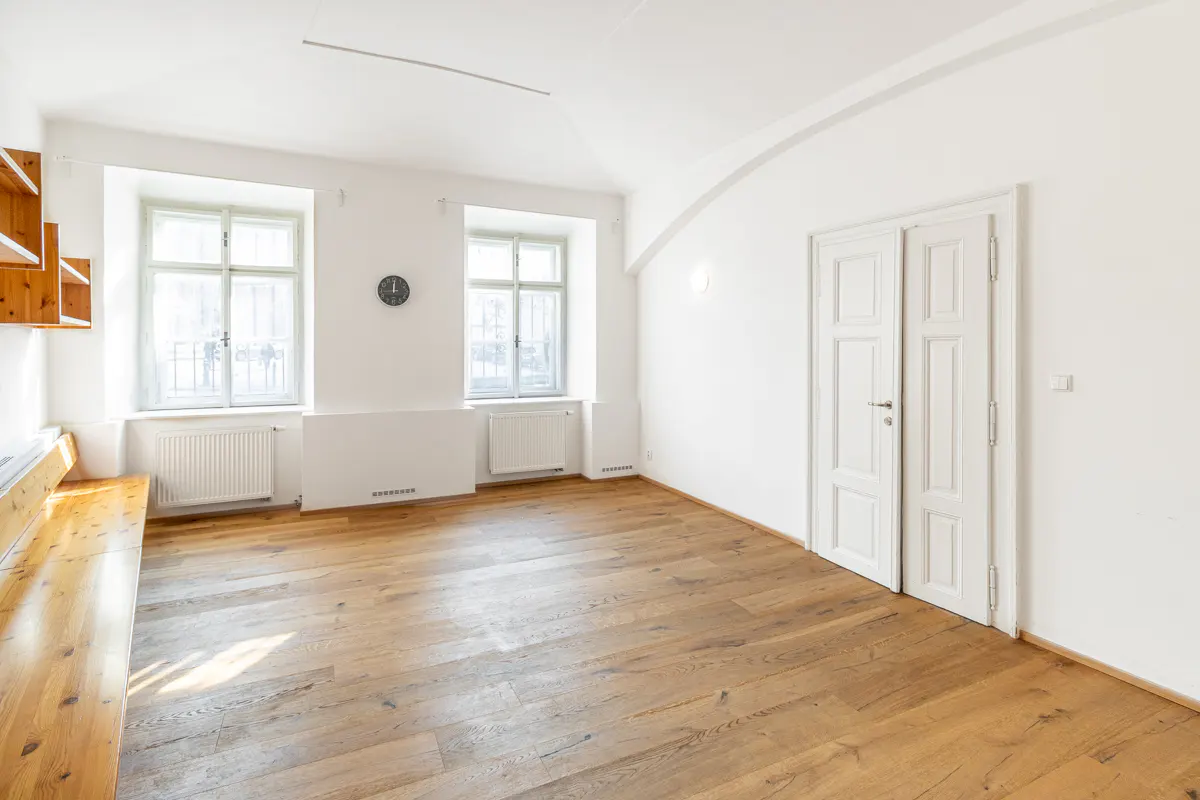 Bright, empty room with hardwood floors, white walls, two windows, and a white double door. A clock hangs on the wall.
