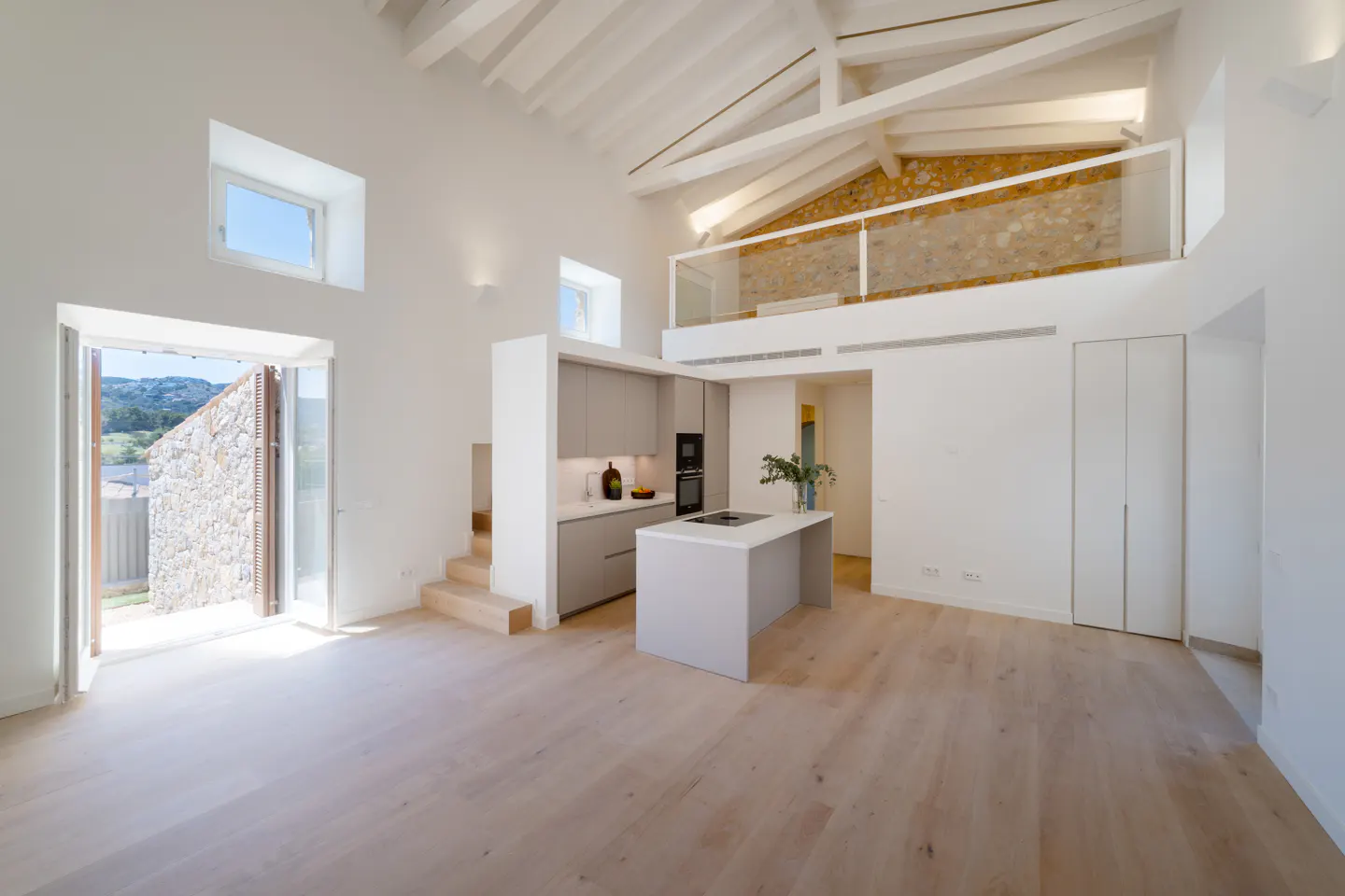 Bright, modern interior with white walls, wood floors, and open kitchen. A stone wall is visible through open doors. A loft overlooks the kitchen.
