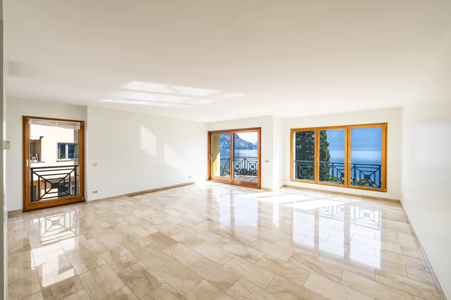 Bright, empty room with marble floors and white walls. Large windows and doors offer a view of the sea and mountains.