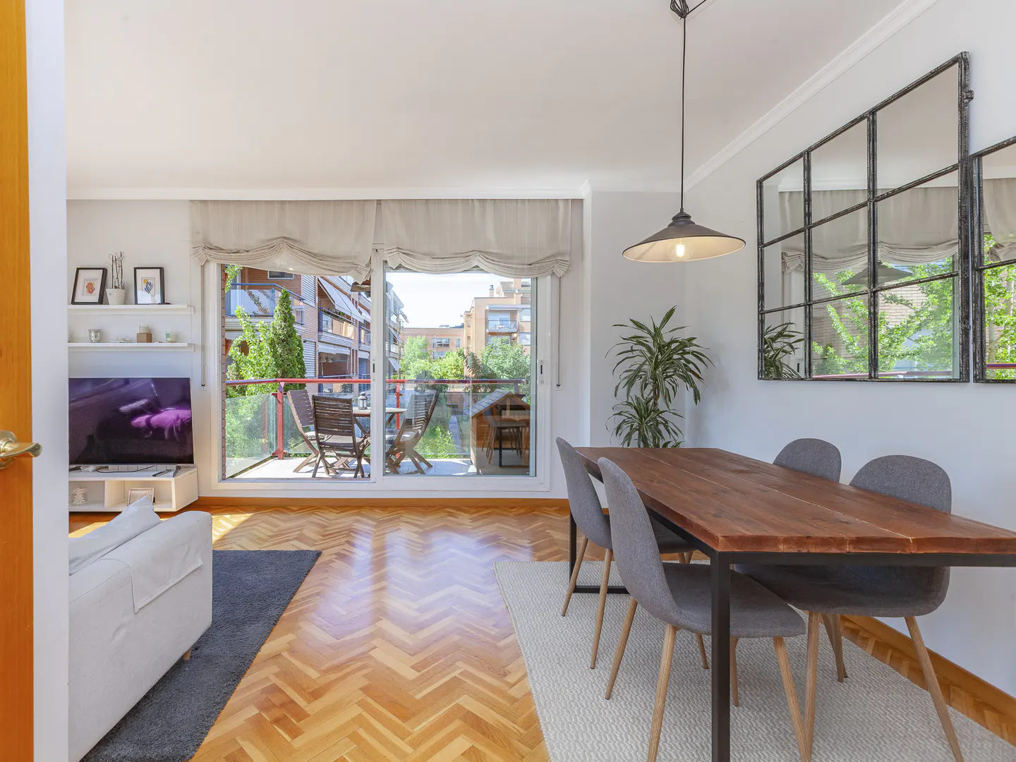 Bright living room with wood floors, dining table with four chairs, and balcony with outdoor seating. Large window and mirror reflect the outside view.