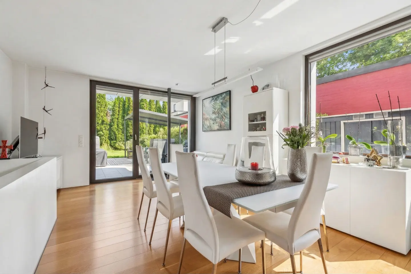 Bright dining room with white chairs, a modern table, hardwood floors, and large windows overlooking a green garden.