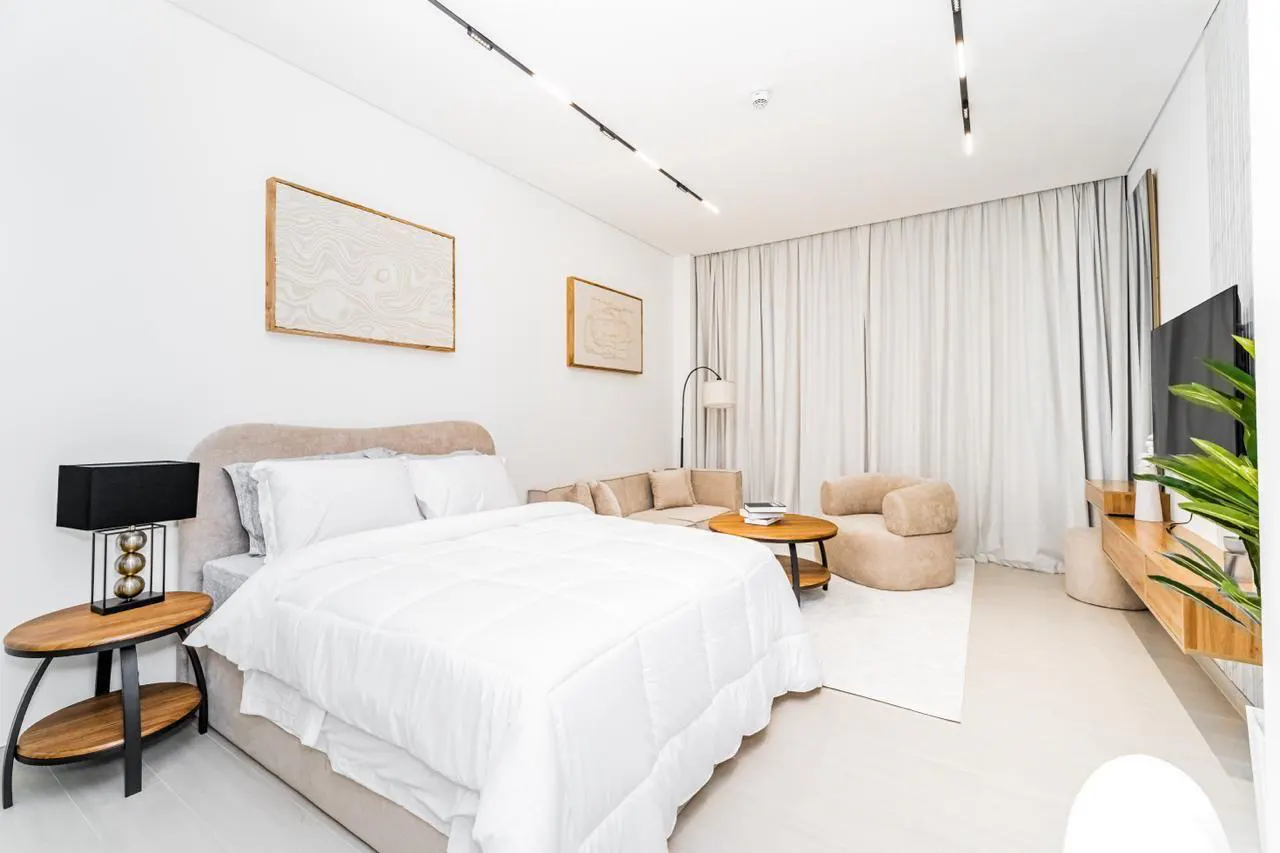 Bright, modern studio apartment with a bed, sofa, chair, and TV. White walls, curtains, and bedding create a clean, minimalist aesthetic.