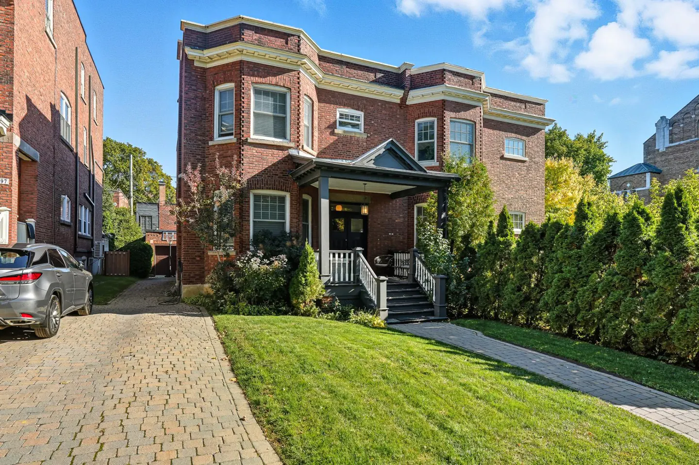 Brick two-story house with a covered porch, green lawn, and a stone driveway. A gray car is parked on the left.