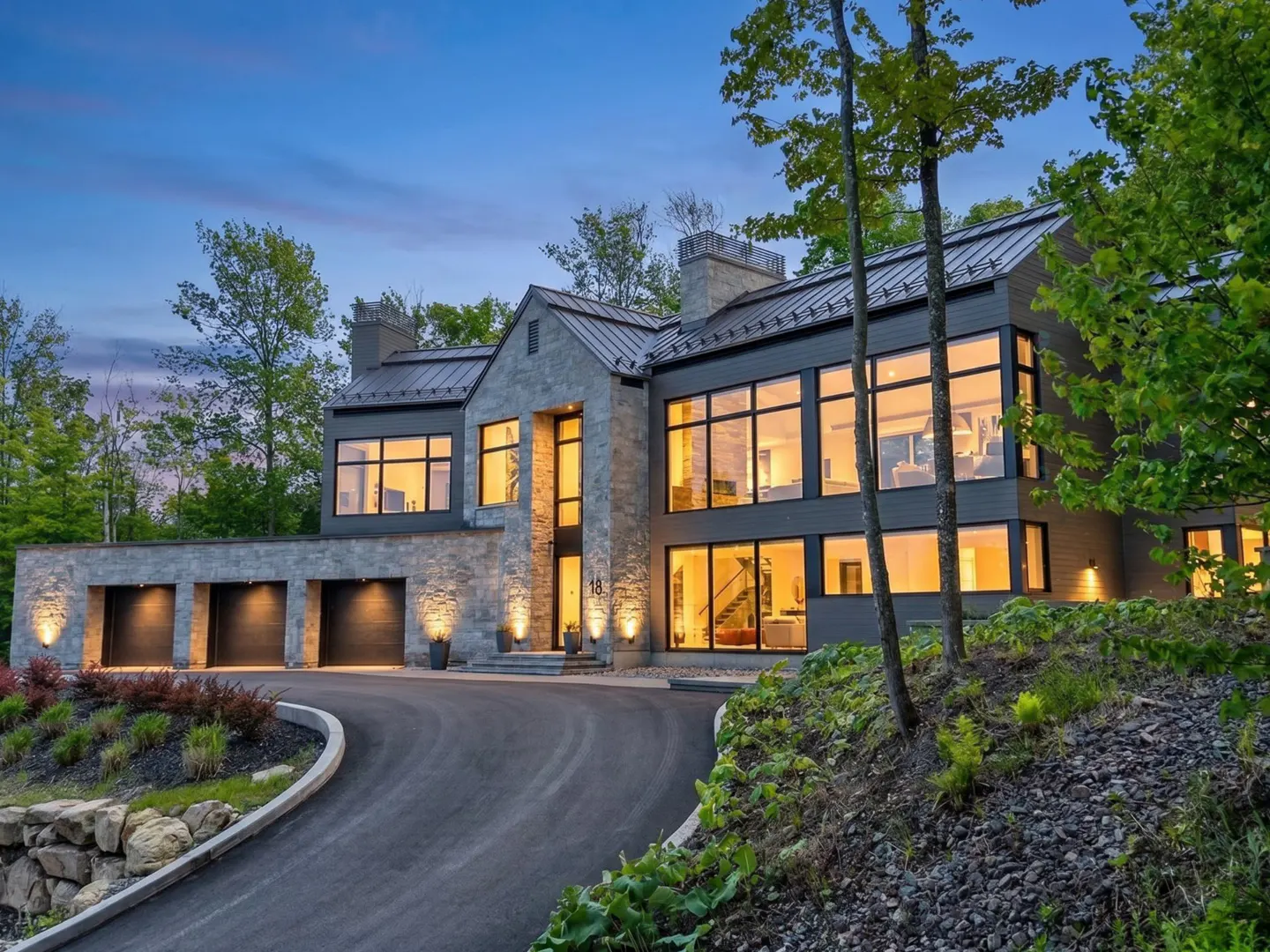 Modern stone and dark metal home at dusk with a curved driveway, illuminated windows, and surrounding lush greenery.