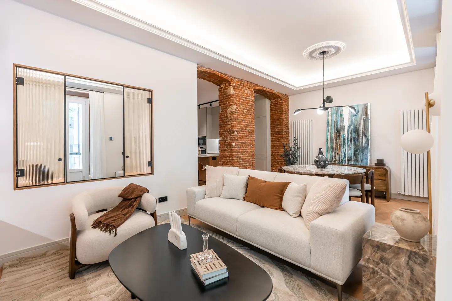 A bright living room with a white sofa, black coffee table, and a mirror. A dining area with a round table is visible in the background.