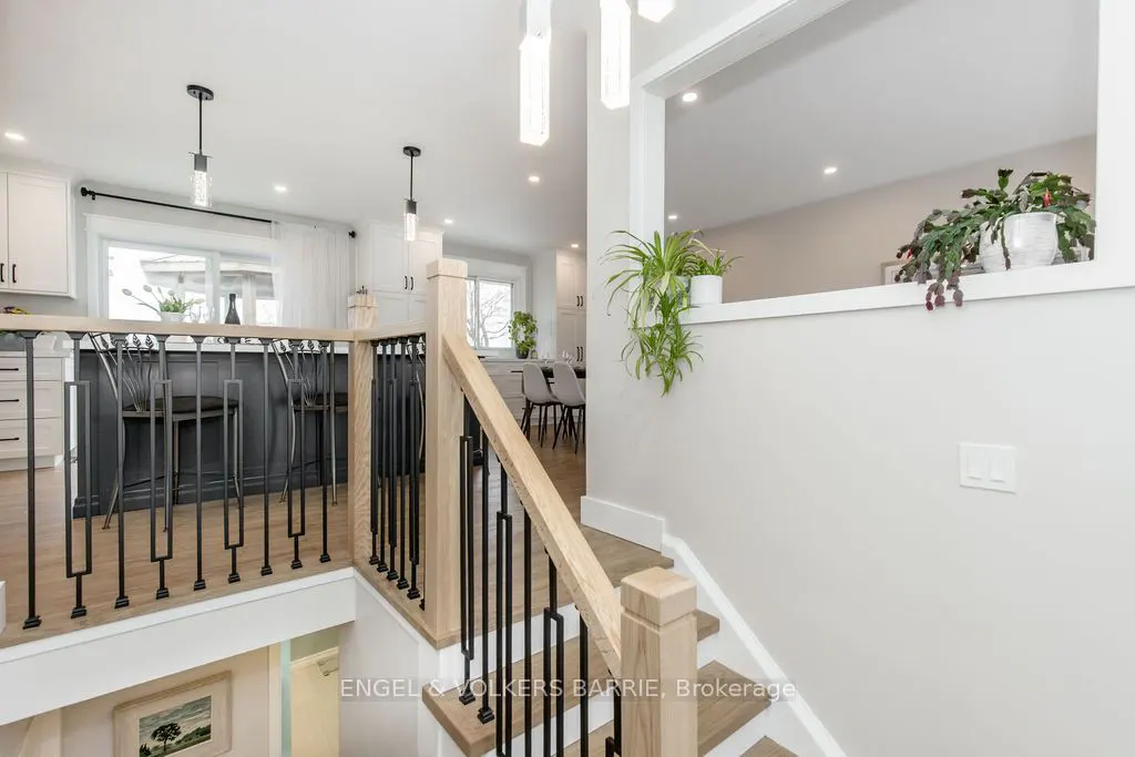 A bright, modern home interior featuring a staircase with light wood and black metal railings, leading to an open-concept kitchen with white cabinets and a dark island. Plants adorn the walls.