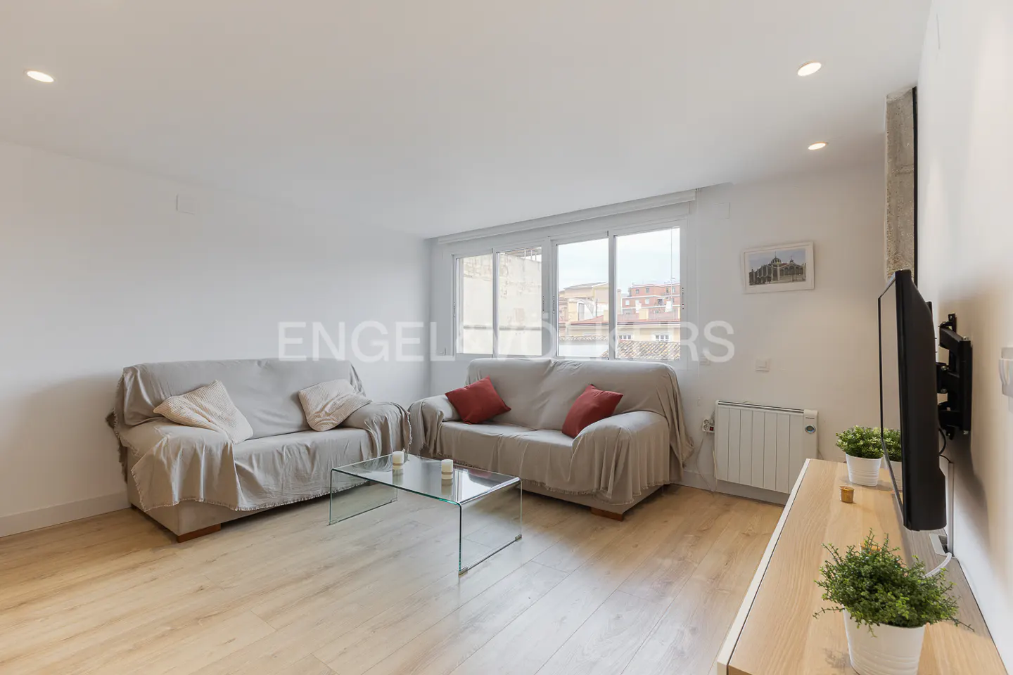Living room with two sofas, glass table, TV, and light wood floors. Pillows and throws add comfort. Natural light from the window.