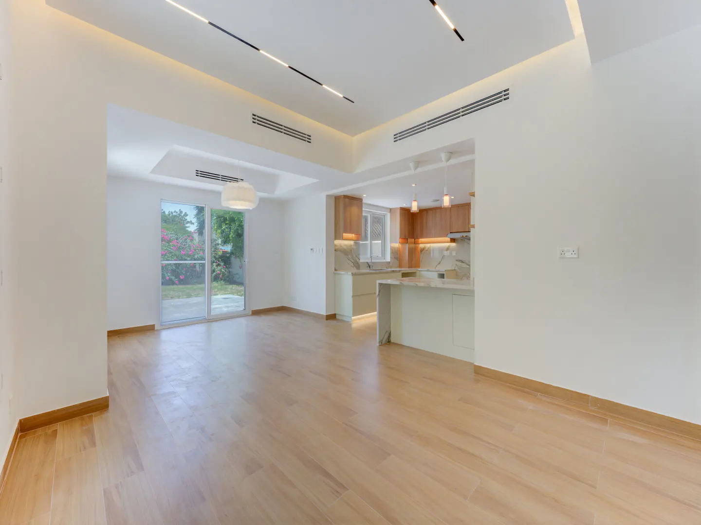 An open-concept living space with light wood floors, white walls, and a modern kitchen with wood cabinets. Sliding glass doors lead to a garden.