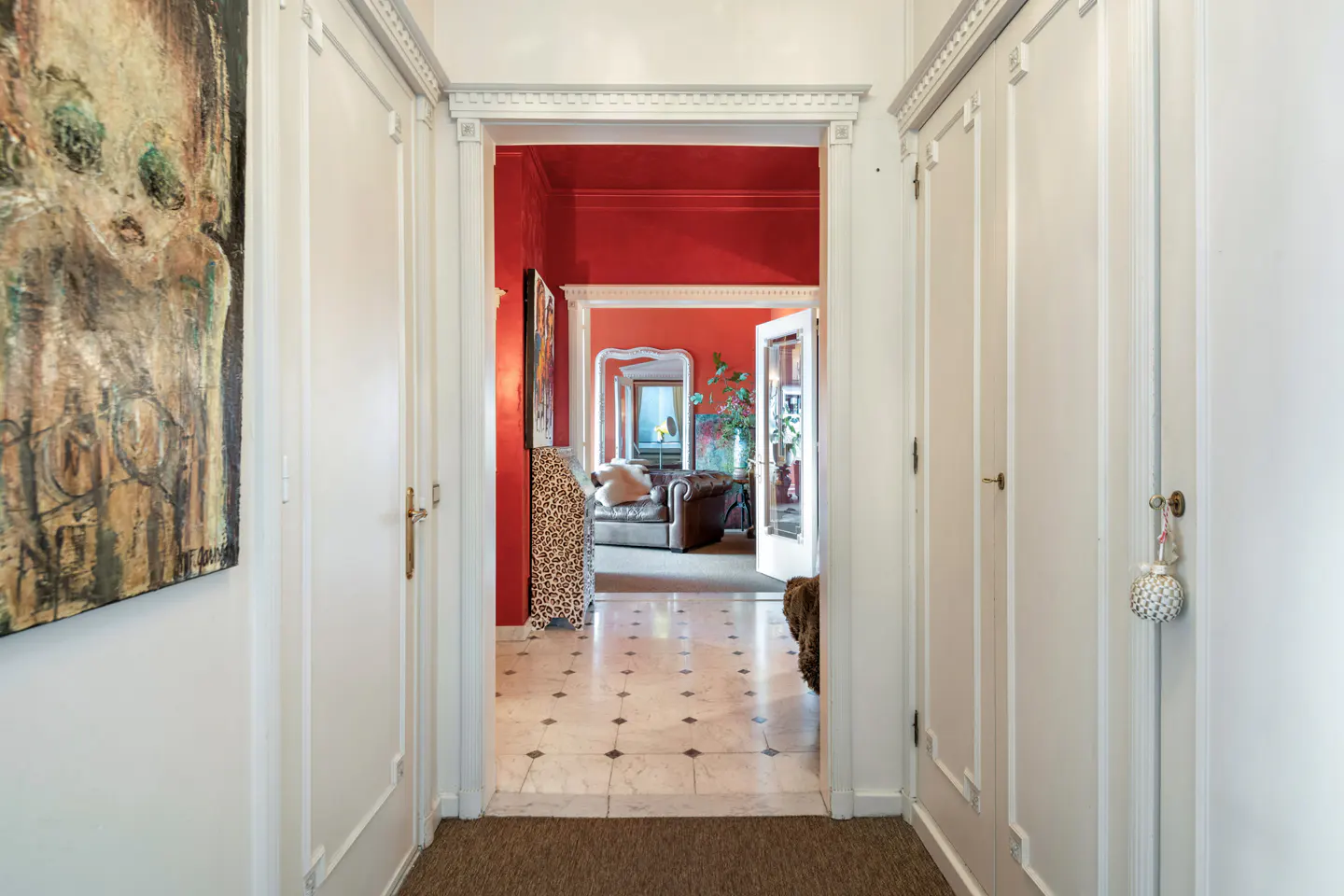 Hallway with white walls, doors, and trim. A red room is visible through the doorway, with a brown leather couch and marble floors. An abstract painting hangs on the left wall.