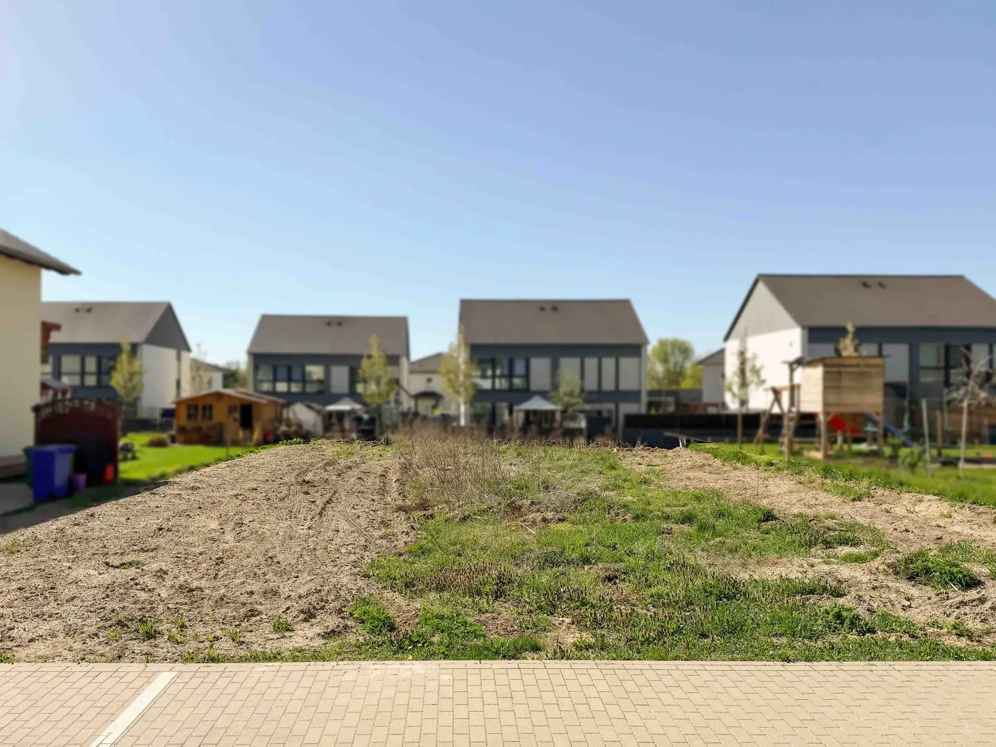 Vacant lot with dirt and grass, ready for building. Modern houses in the background under a clear blue sky.