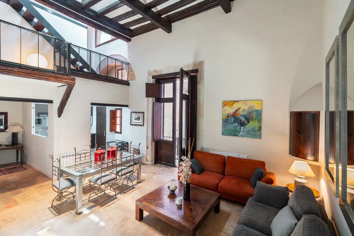 High-ceiling living room with terracotta floors, orange and grey sofas, a wooden coffee table, and a metal dining set.