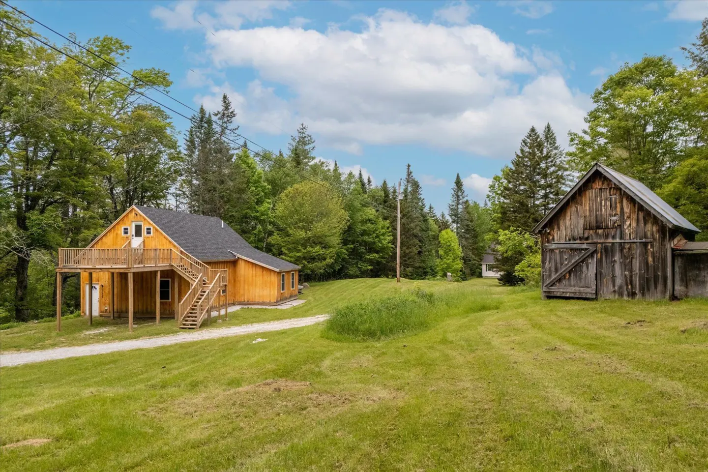 Wooden house with a deck and stairs next to a weathered barn on a green lawn under a blue sky.