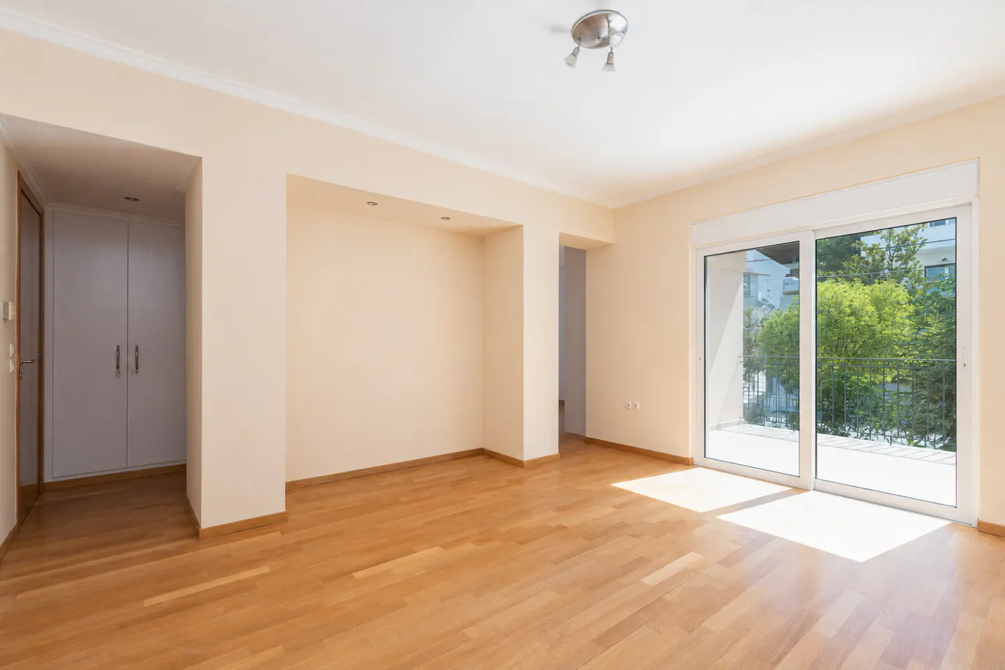 Bright, empty room with light wood floors, cream walls, and white trim. Sliding glass doors lead to a balcony with green trees visible.